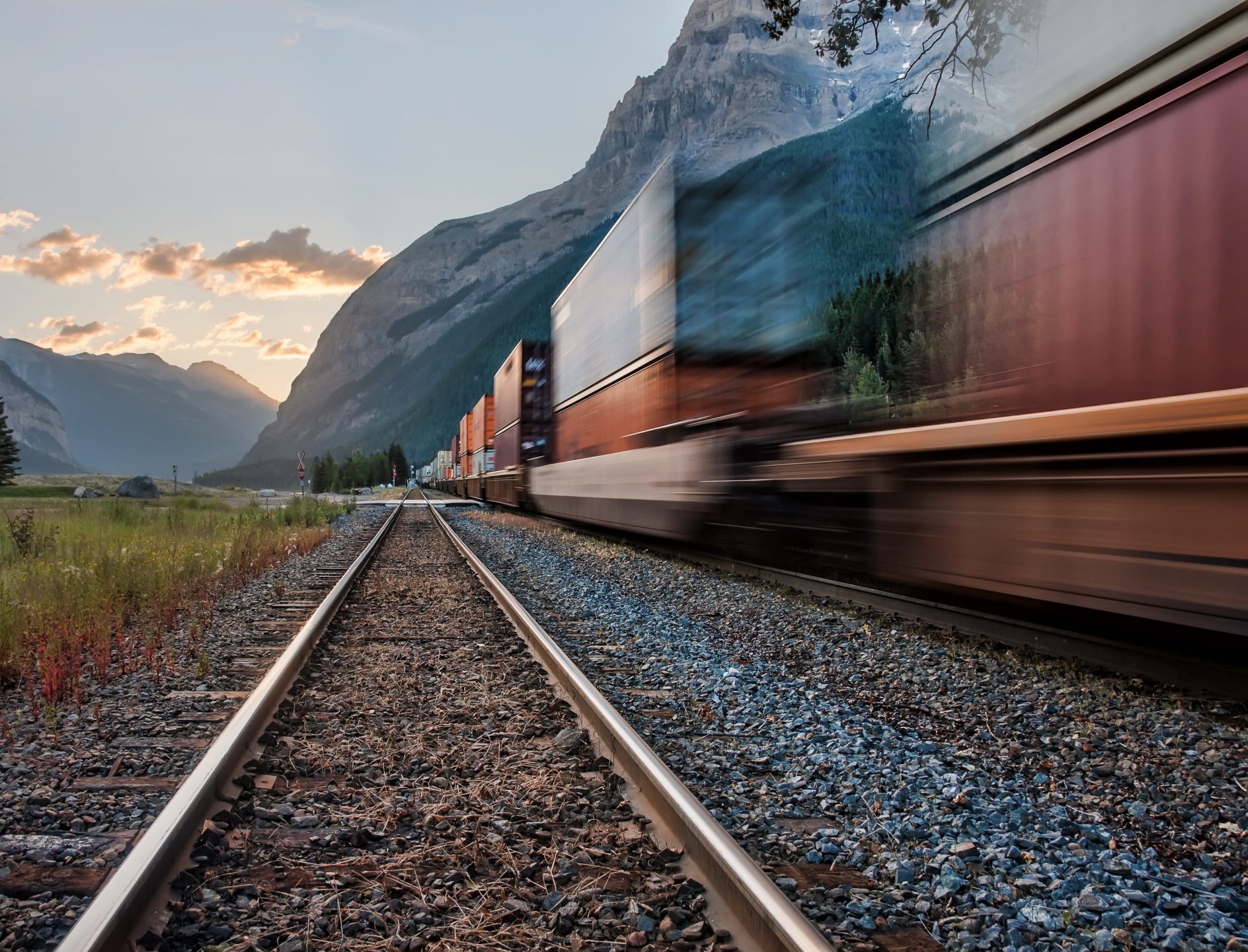 Freight train carrying containers speeding past a mountain valley at dusk
