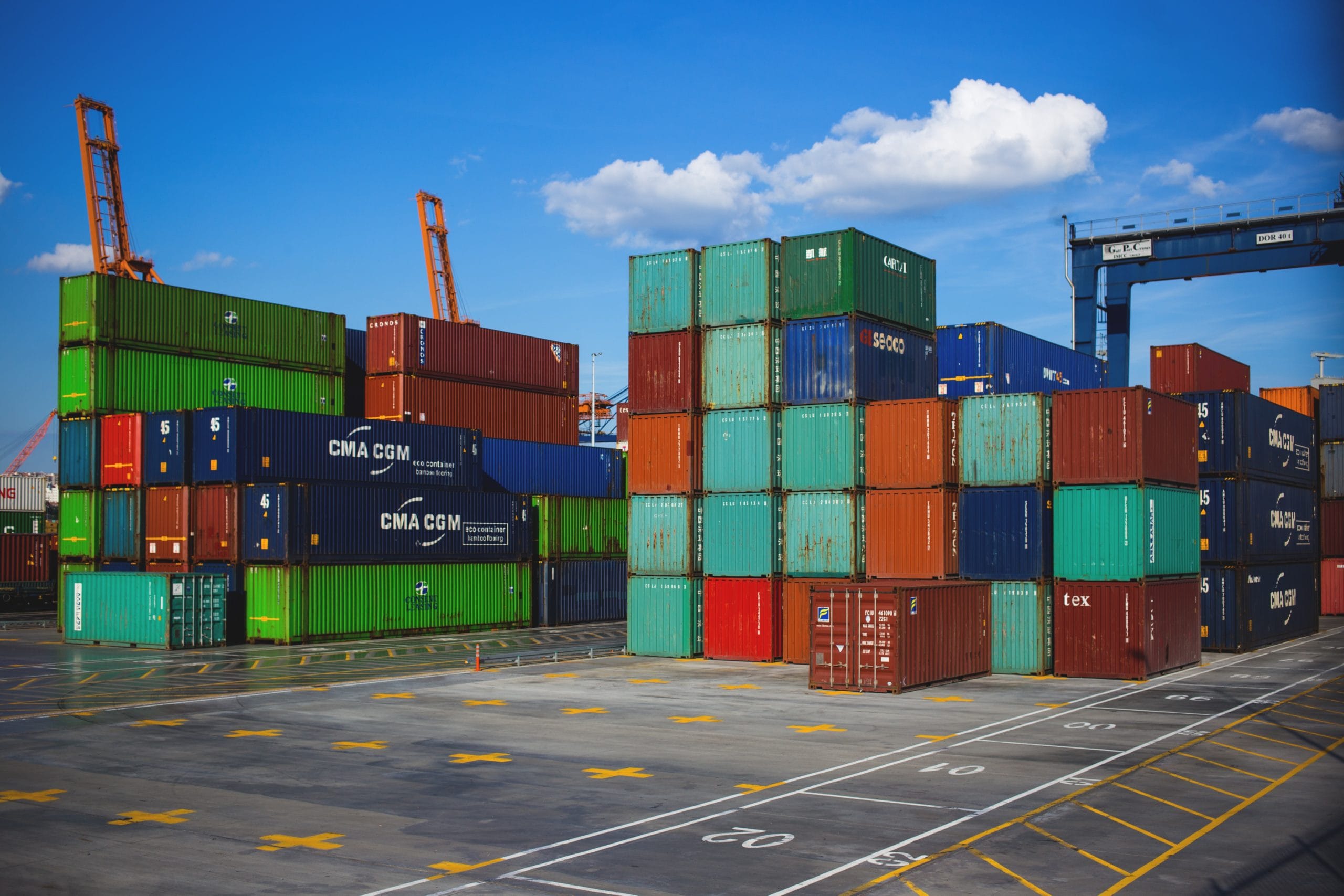 Colourful shipping containers stacked in a port yard with orange gantry cranes under a blue sky