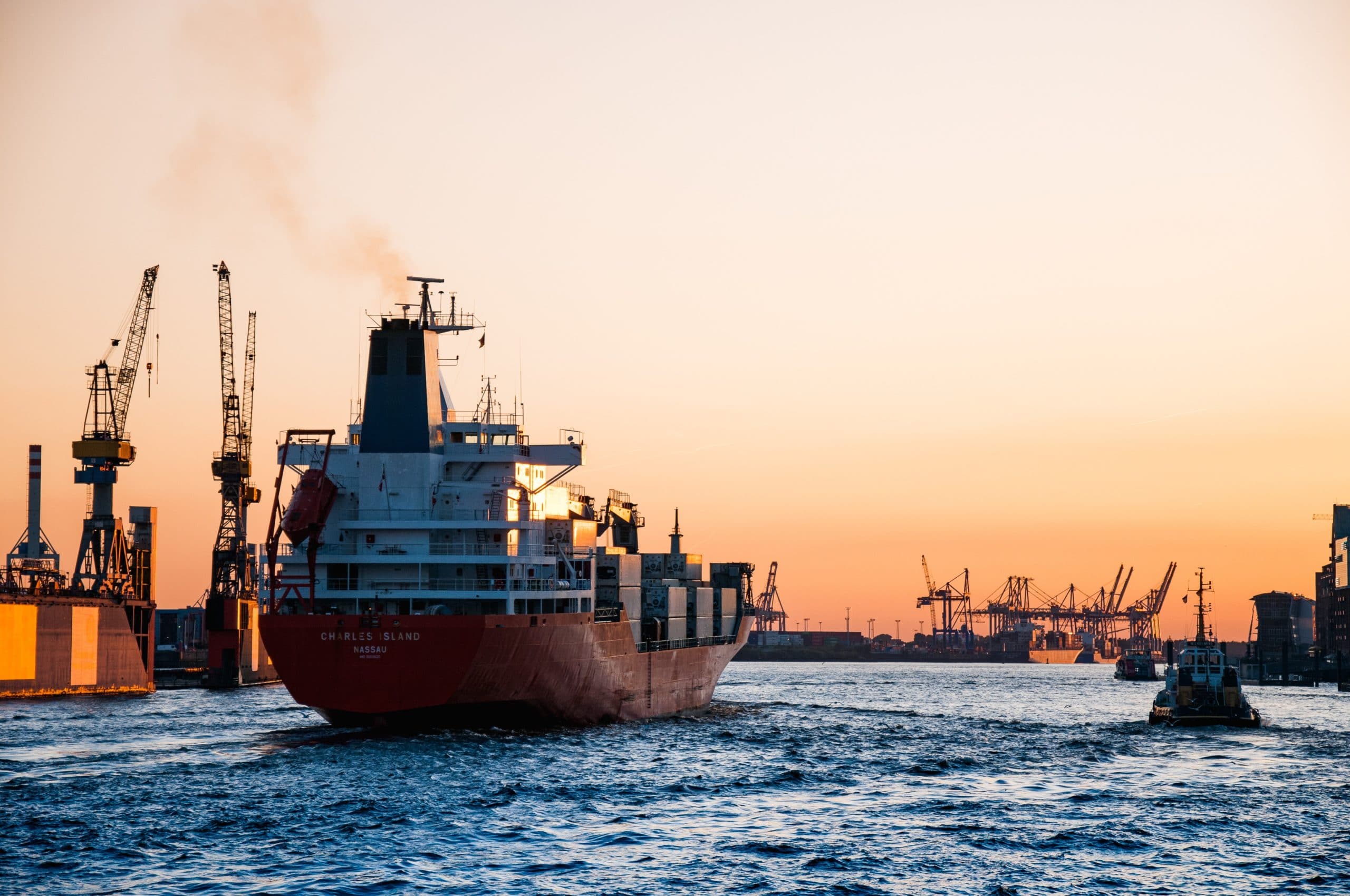 Cargo ship departing a busy port at sunset with cranes silhouetted against an orange sky