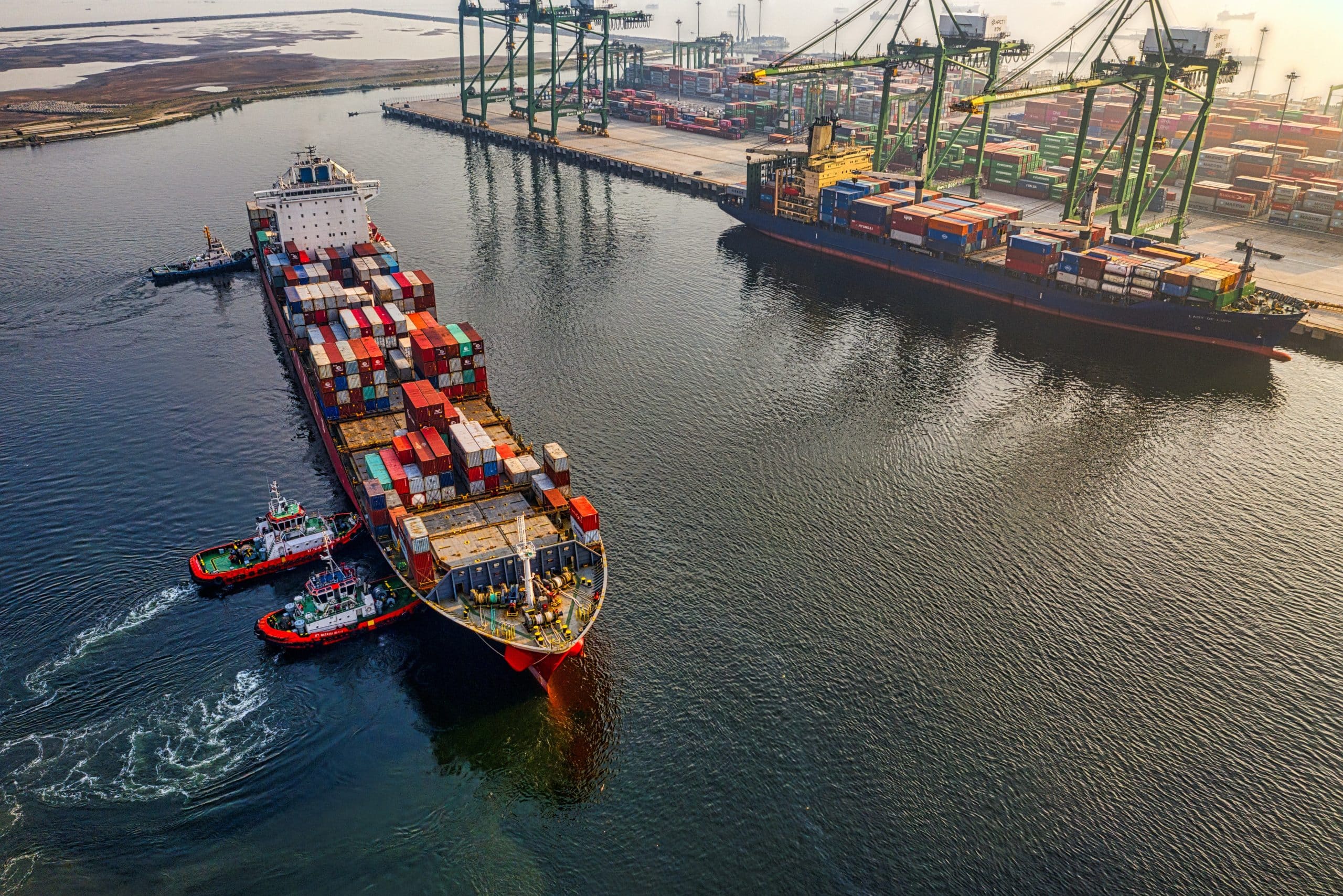 Aerial view of a loaded container ship being guided into port by tugboats with gantry cranes alongside