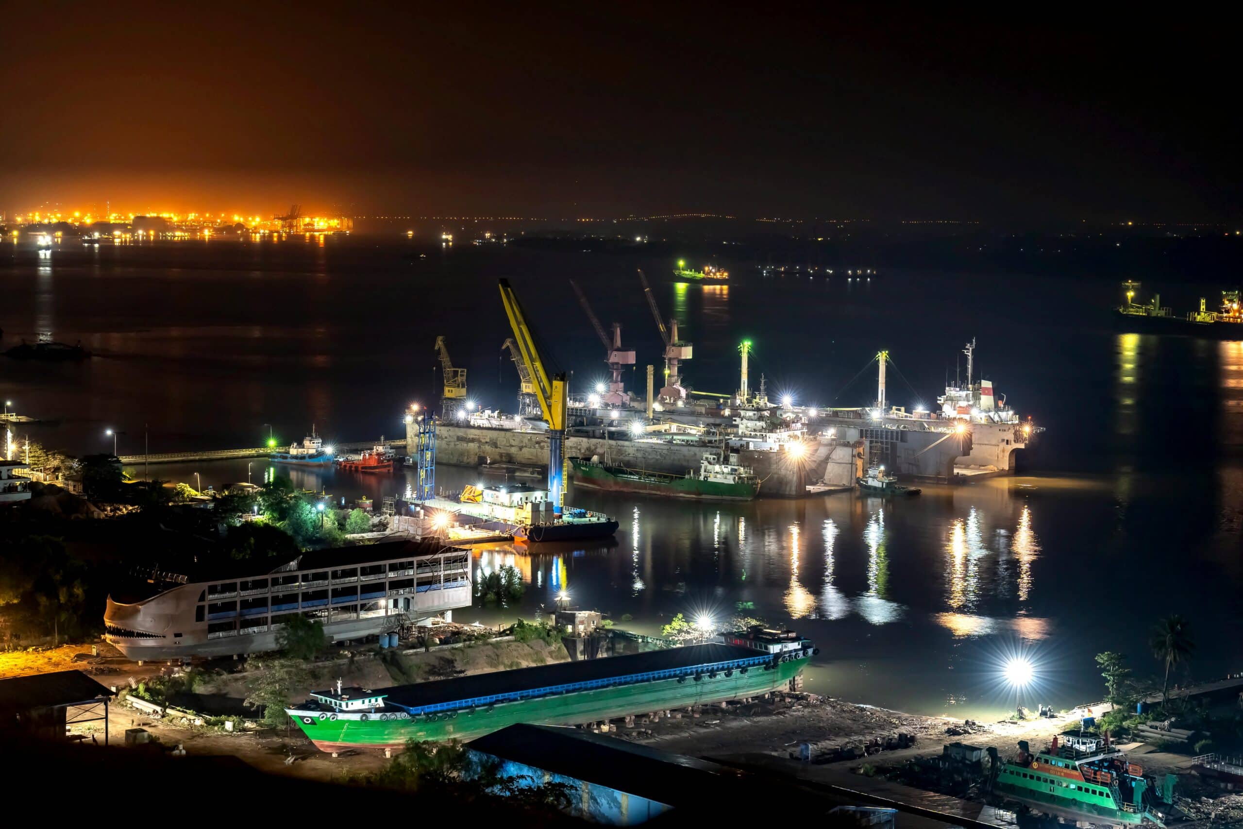 Aerial night view of a busy port and shipyard with illuminated cranes and vessels reflected on the water
